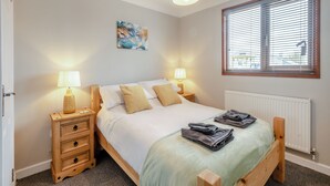 A bedroom with a bed, two bedside tables, a lamp, a radiator, a window with blinds, and a painting on the wall.