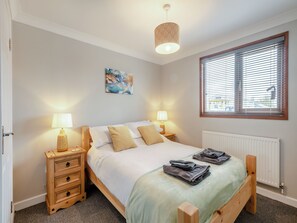 A bedroom with a bed, two bedside tables, a lamp, a radiator, a window with blinds, and a painting on the wall. - Perfect riverside holiday accommodation, set on the water’s edge at Brundall. (Brundall)