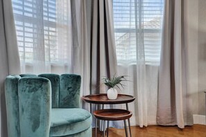 Quarto com uma poltrona estofada em tom de verde-água, uma pequena mesa de madeira com uma planta e cortinas translúcidas.