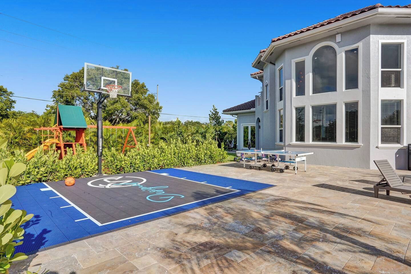 A basketball court with a hoop, a swing set, and a house with large windows.