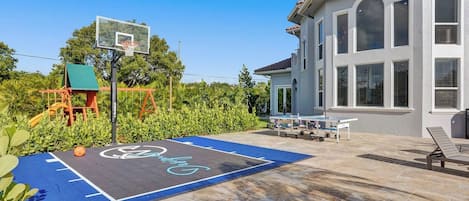 A basketball court with a hoop, a swing set, and a house with large windows.
