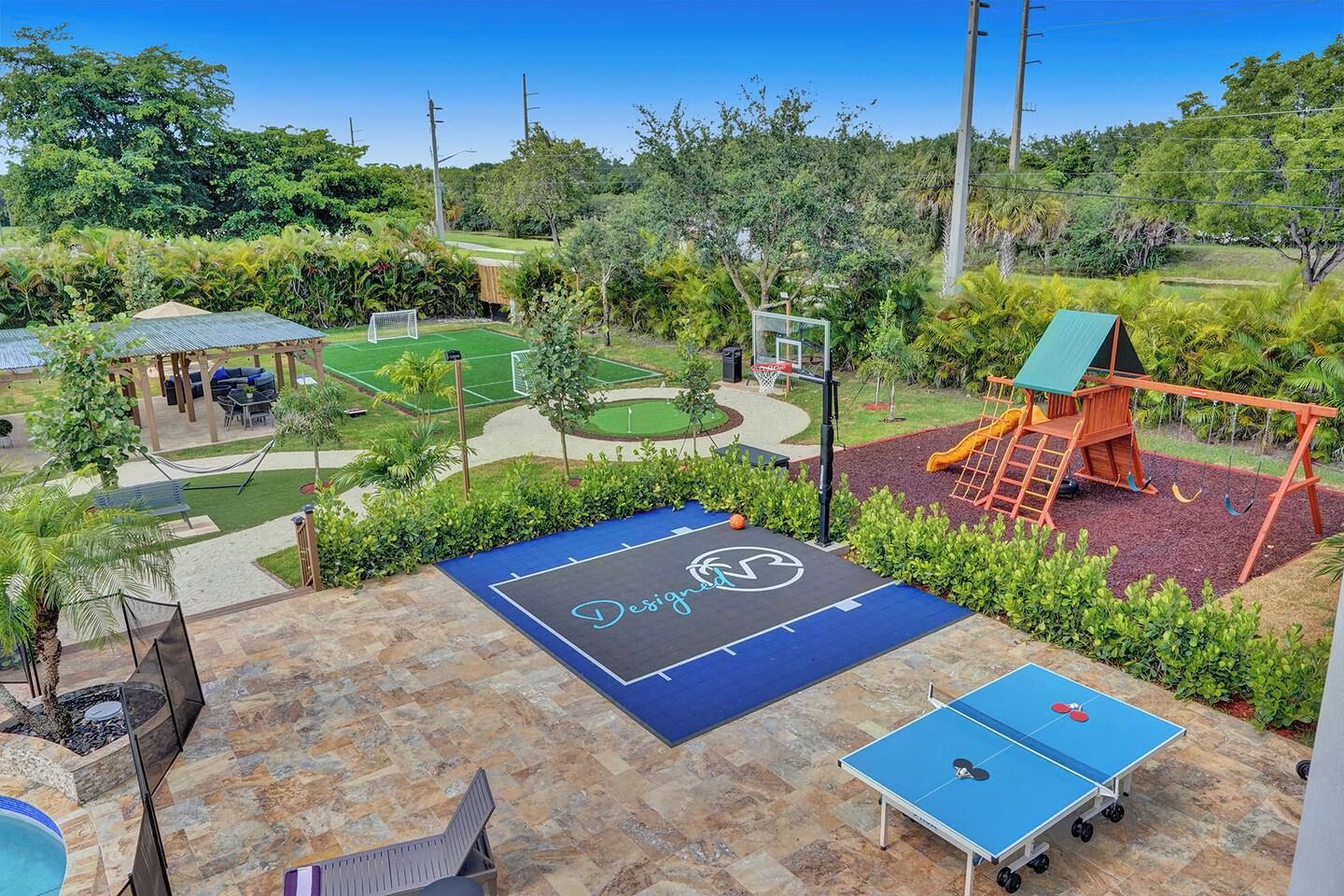 A backyard with a basketball court, a ping pong table, a swing set, and a covered seating area.