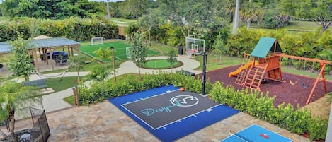 A backyard with a basketball court, a ping pong table, a swing set, and a covered seating area.