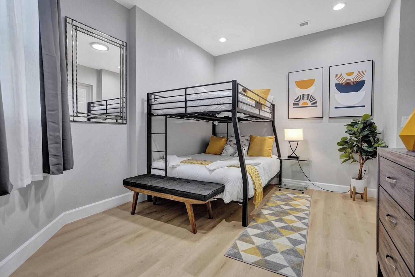 Un letto a castello con un comodino e una lampada, una piccola pianta e un comò in una stanza moderna con un tappeto a motivi geometrici.