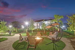 Un espace extérieur aménagé avec des chaises et un foyer, entouré de pelouses et d’arbres bien entretenus, sous un ciel crépusculaire.