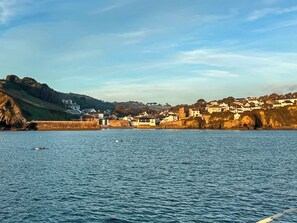 Une ville côtière avec des bâtiments le long de la plage, une étendue d’eau au premier plan et un paysage vallonné en arrière-plan.