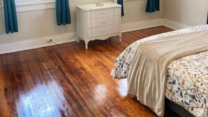 Une chambre avec un plancher en bois, un lit recouvert d’une housse à motifs, une table de chevet avec une lampe et deux fenêtres avec des rideaux.