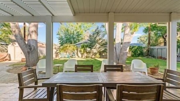 Een overdekt terras met een houten tafel en stoelen, omgeven door een tuin met bomen en gazon.