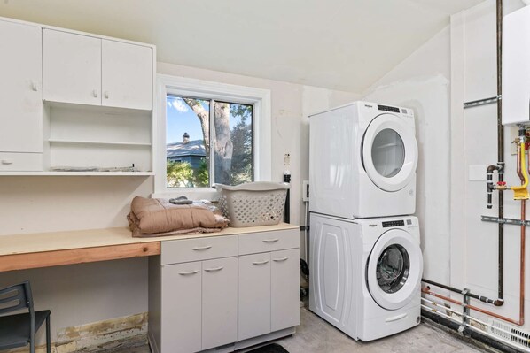 A laundry room with a stacked washer and dryer, a window with a view of trees, and a wooden table with a cushion. - Reno Roost a welcoming private home with yard in Old SW Reno (Reno)