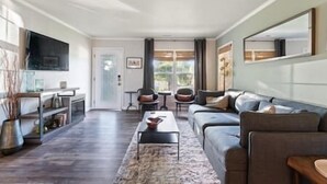 Een moderne woonkamer met een bank, salontafel en stoelen. Er is een televisie aan de muur bevestigd en een deur die naar een andere kamer leidt.