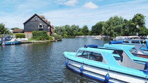 A row of blue boats docked at a calm river with a house and trees in the background. - Watchkeeper's Cottage (Mundesley)