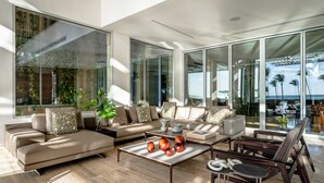 Een moderne woonkamer met een beige bank, een salontafel met decoratieve objecten en uitzicht op de oceaan door grote glasramen.