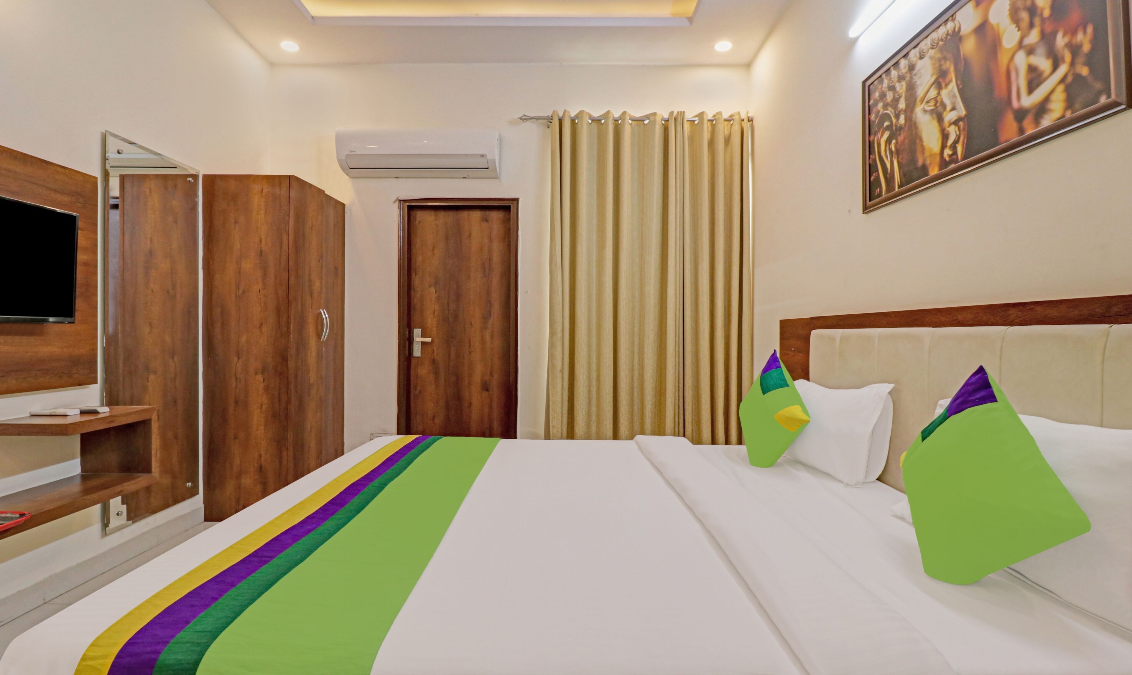 A hotel room with a bed, a TV, wooden wardrobe, and a painting on the wall.