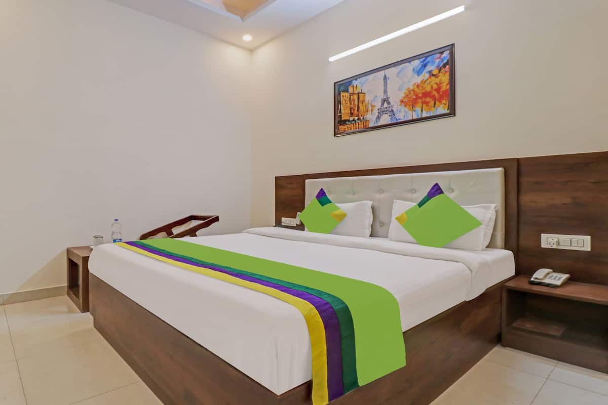 A hotel room with a large bed, a bedside table with a phone, a water bottle, and a painting of the Eiffel Tower.