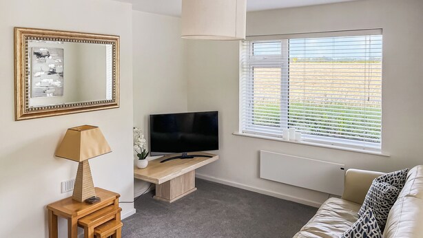 A modern living room with a flat-screen TV, a wooden side table, a lamp, a beige sofa, and a large window with blinds. - 2 bedroom accommodation in Caythorpe, near Nottingham (Caythorpe, near Nottingham)