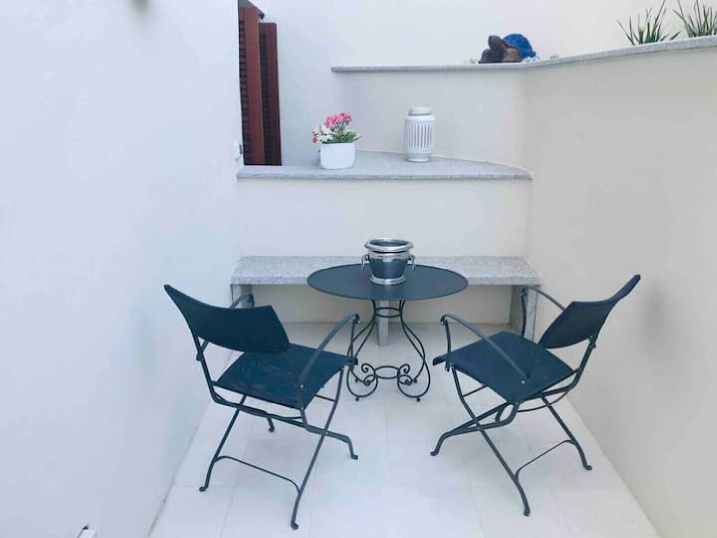 A small outdoor patio with a round table and two chairs, a potted plant, and a white lamp on a shelf.