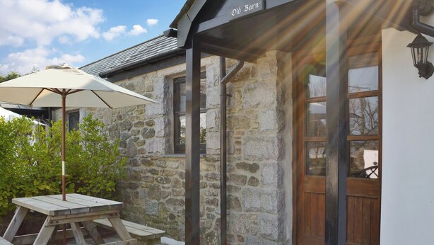 A stone building with a wooden door and a covered patio area with a table and umbrella. - Old Barn Cottage - UK11089 (St Breward, near Bodmin)