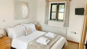 A neatly made bed with white linens, a bedside table with a lamp, a decorative wall piece, and a window with curtains. - Old Barn Cottage - UK11089 (St Breward, near Bodmin)