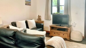 A living room with a black leather sofa, a wooden TV stand, a flat-screen TV, a small wooden side table, and a window with curtains. - Old Barn Cottage - UK11089 (St Breward, near Bodmin)