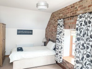 A bedroom with a bed, a radiator, a lamp, a wooden wardrobe, a brick wall, and a window with floral curtains. - 2 bedroom accommodation in North Somercotes, Mabelthorpe (North Somercotes, Mabelthorpe)