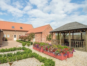 A brick building with a red-tiled roof, a garden with flower beds, and a wooden pavilion with outdoor seating.
