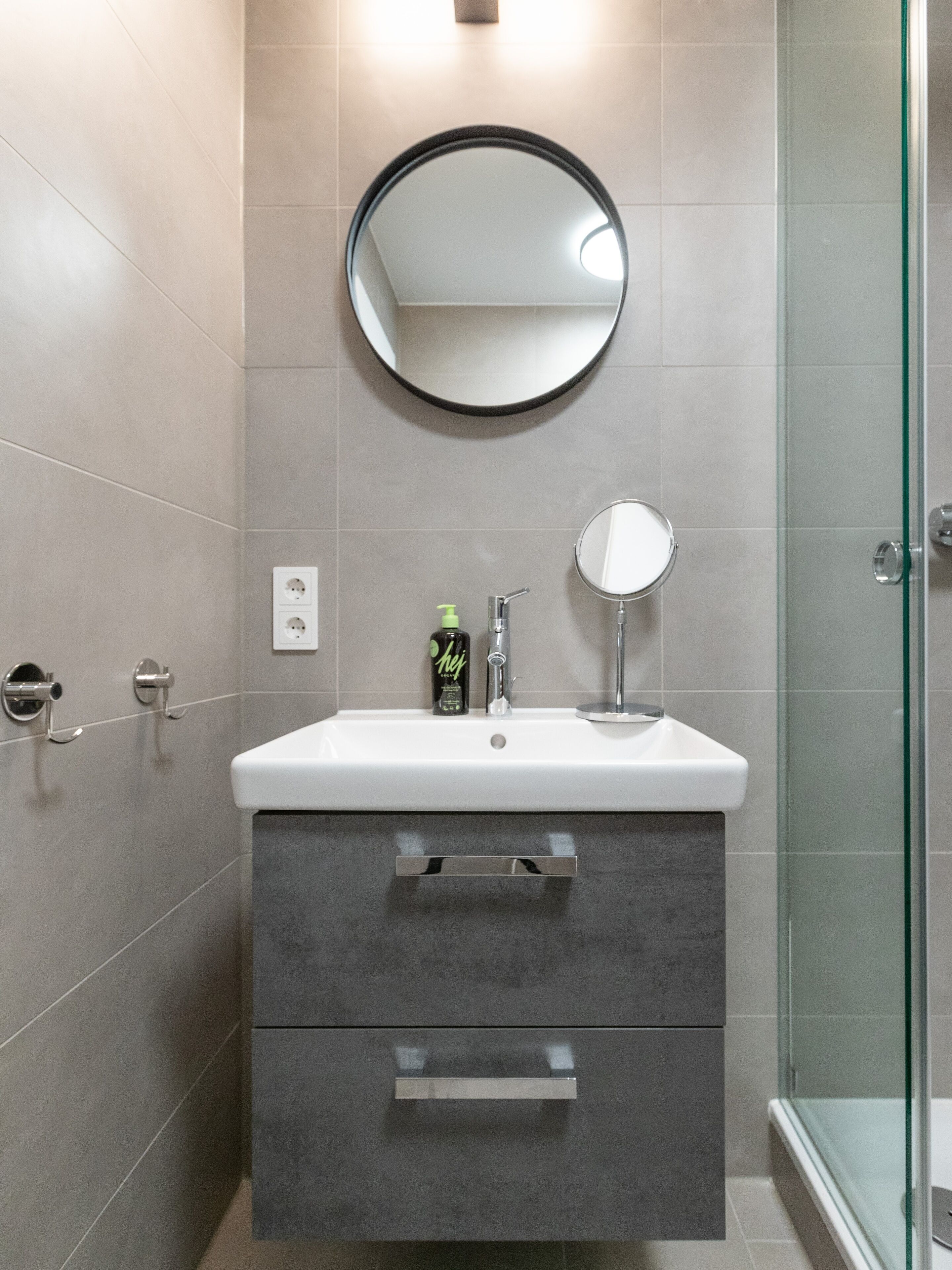 Un baño moderno con un lavabo blanco, dos cajones, un espejo redondo y una cabina de ducha de vidrio.