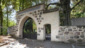 Um arco de pedra com um portão de metal, rodeado por árvores e um muro de pedra.