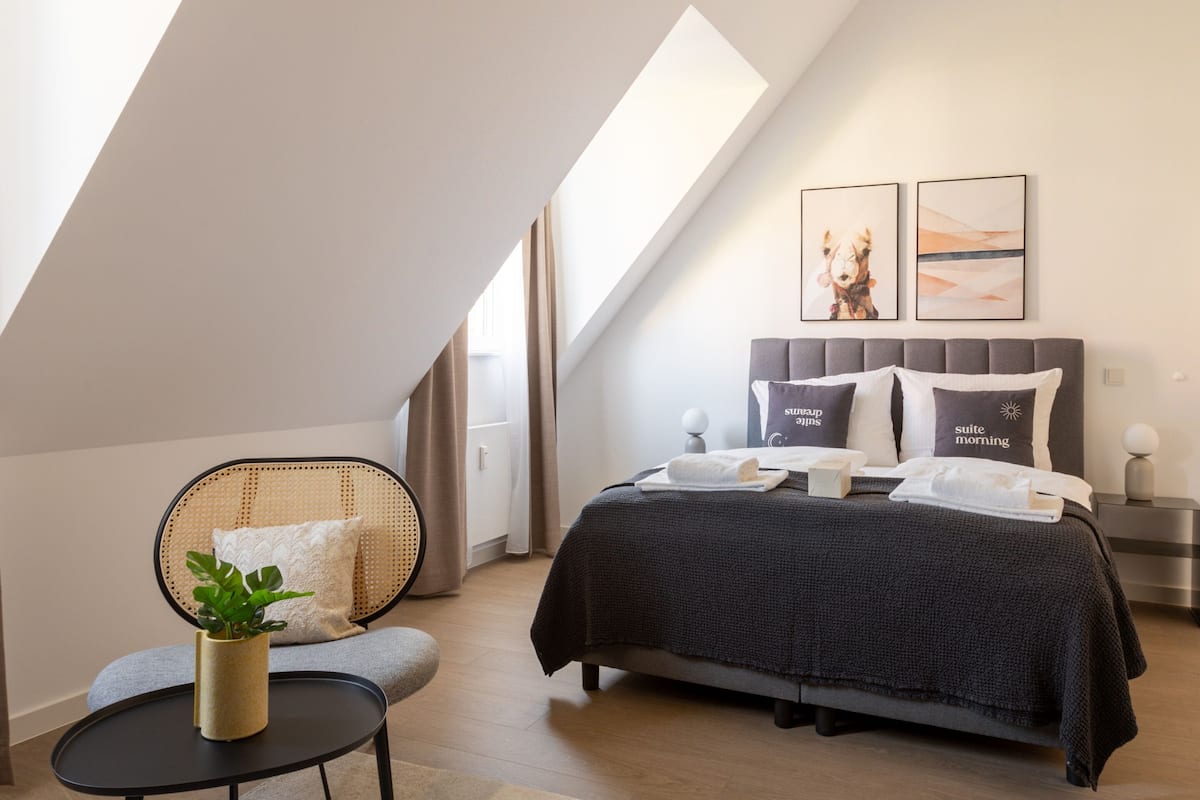 Une chambre moderne avec un plafond en pente, un lit recouvert d’une couverture sombre, une table de chevet avec une lampe et une chaise agrémentée d’une plante.