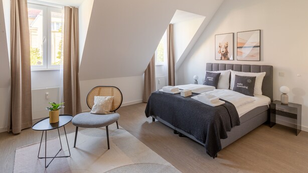 Quarto moderno com uma cama grande, uma cadeira, uma mesinha e uma janela com cortinas.
