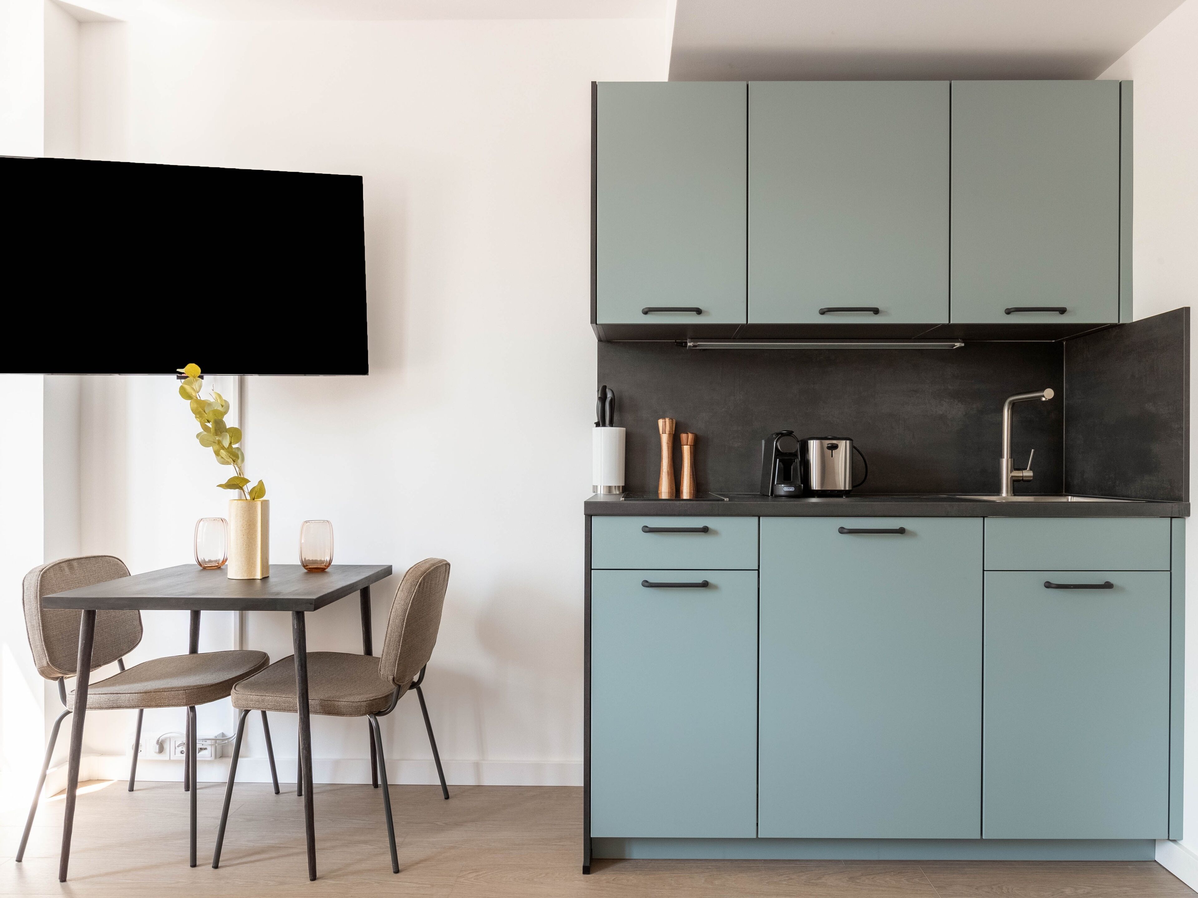 Eine moderne Küche mit hellblauen Schränken, einer schwarzen Arbeitsfläche und einem Essbereich mit zwei Stühlen und einem Tisch.