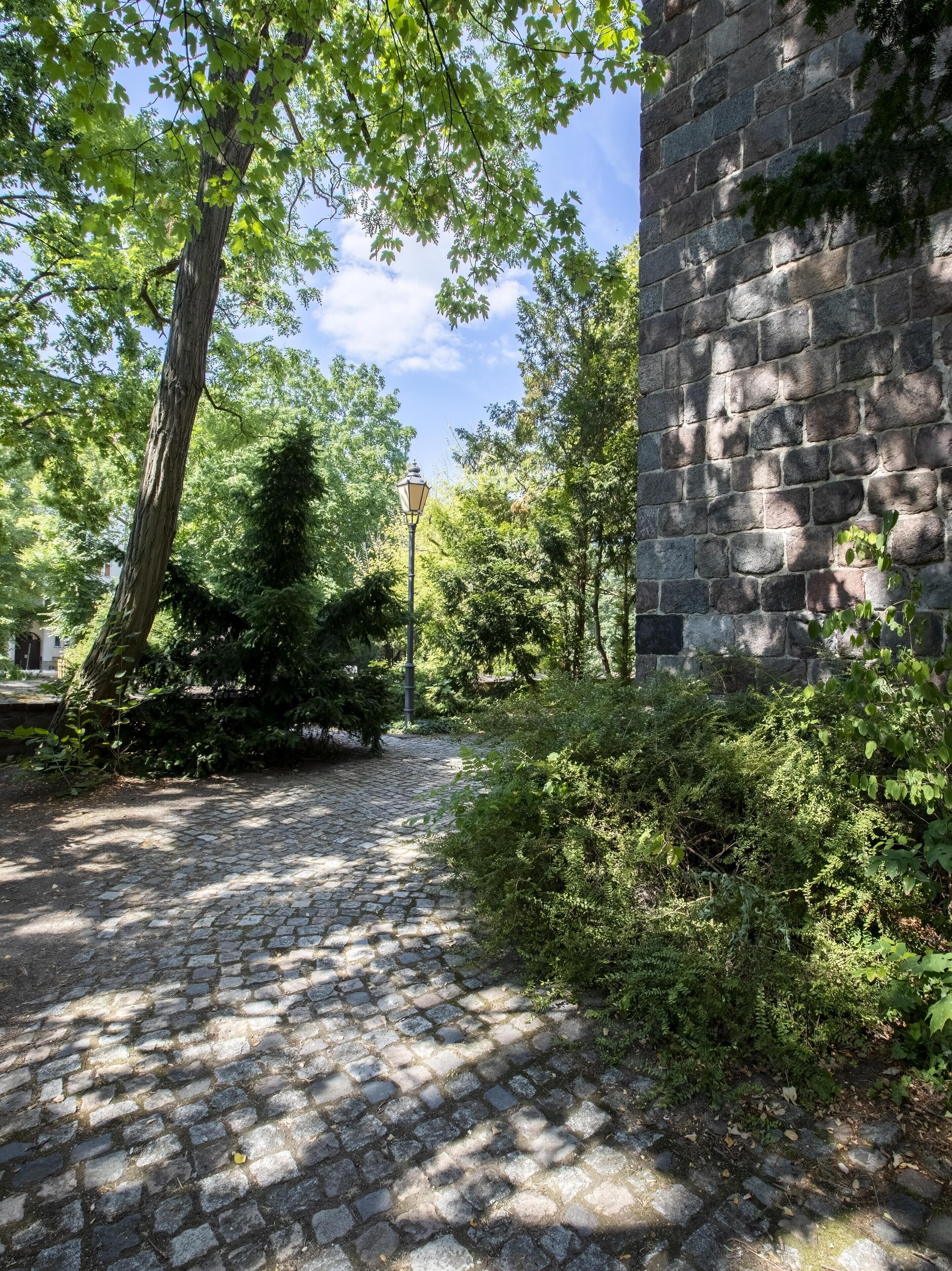 A cobblestone pathway leading to a stone building with a brick wall and a street lamp.
