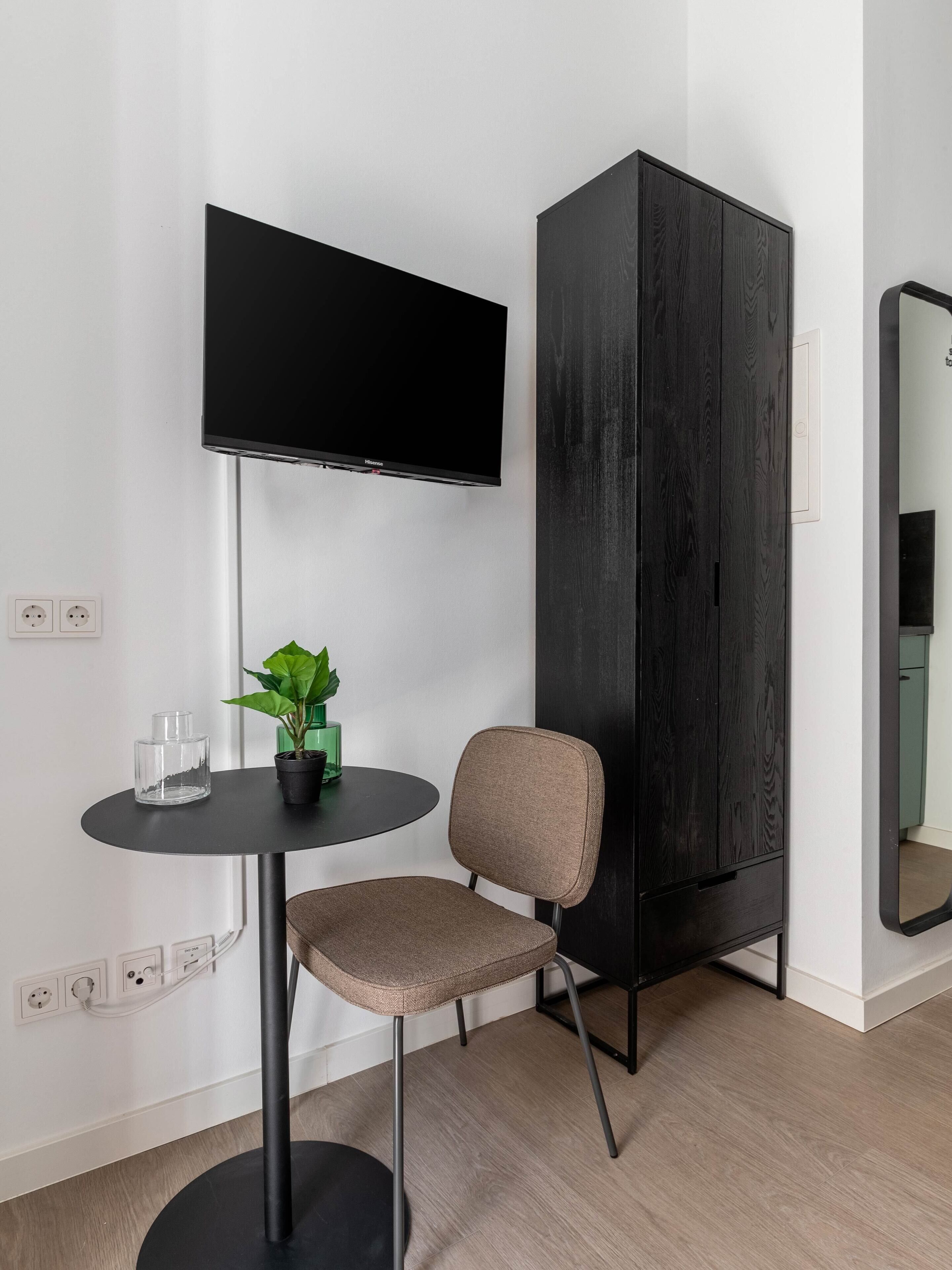 A modern room with a flat-screen TV mounted on the wall, a small round table with a chair, a potted plant, and a dark wooden cabinet.