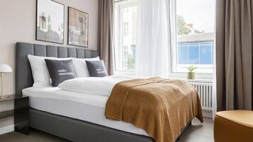 Ett modernt sovrum med en grå, klädd sänggavel, vita sängkläder och en brun pläd. På väggen hänger två inramade tavlor, och det finns en sänglampa och ett fönster med gardiner.