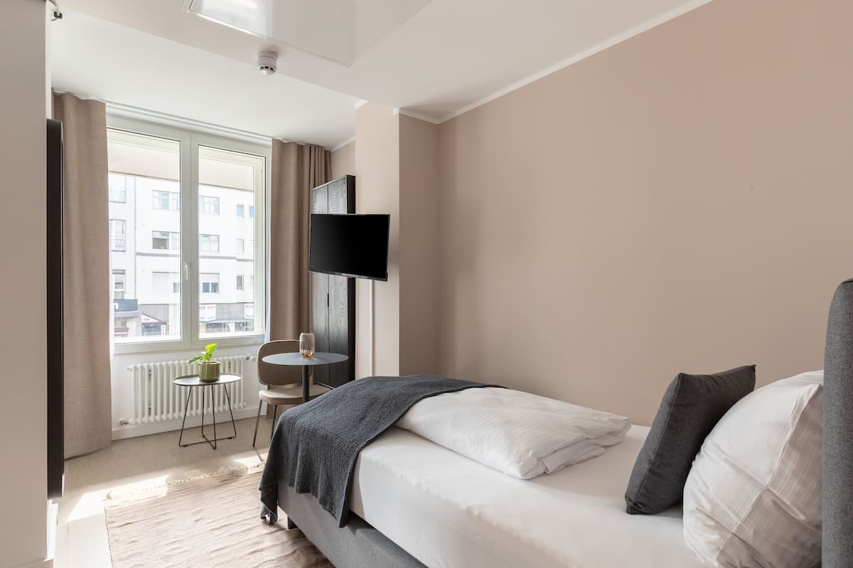 Une chambre d’hôtel moderne avec un grand lit, une petite table ronde avec une chaise, un téléviseur à écran plat fixé au mur et une fenêtre avec des rideaux.