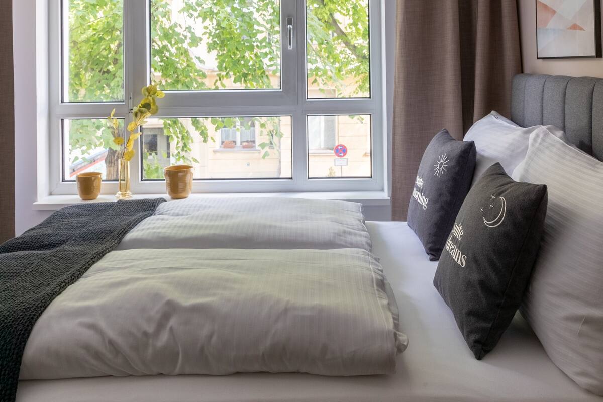 Une chambre avec une grande fenêtre, un lit avec des oreillers et un vase rempli de fleurs.