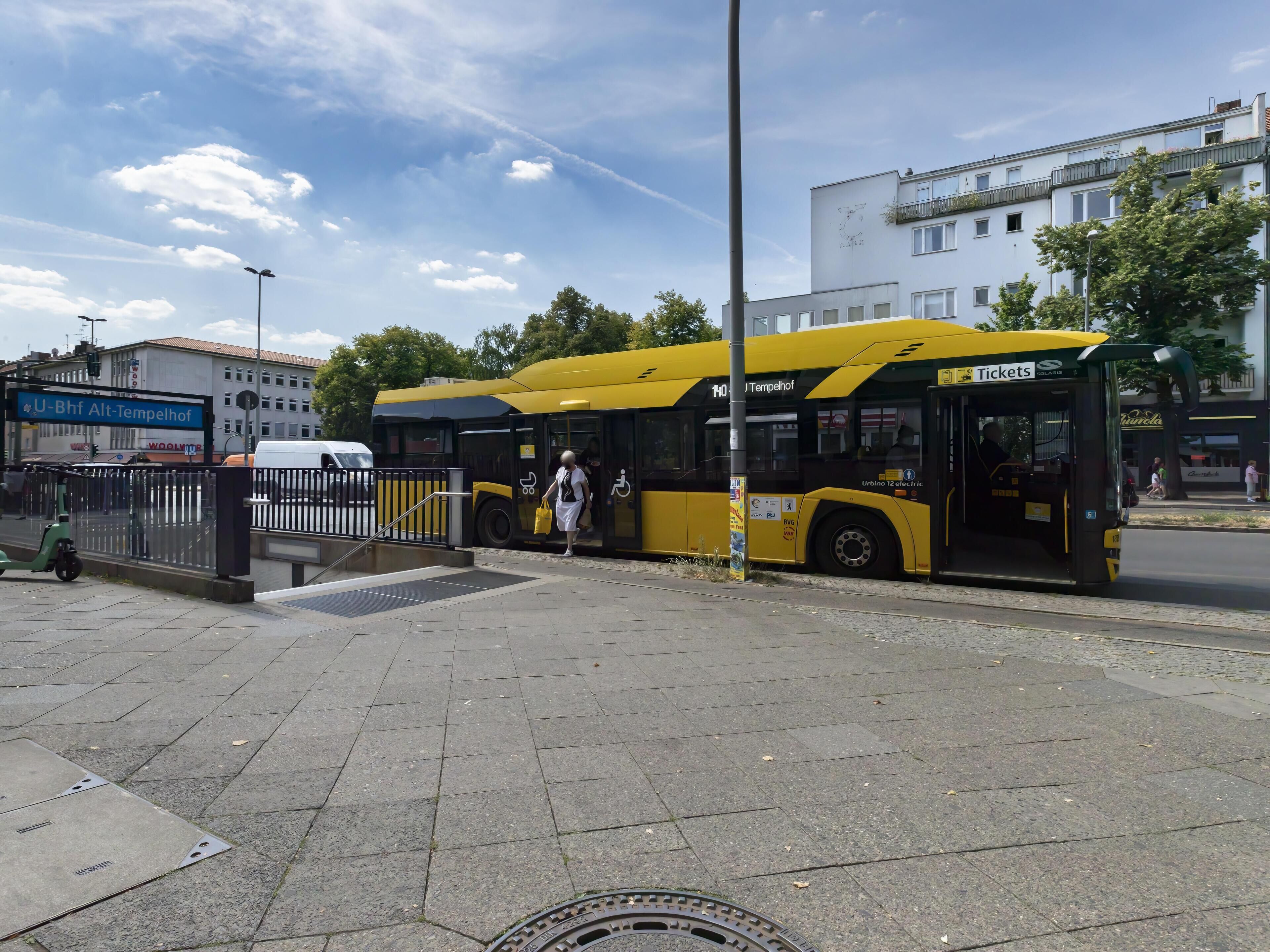 A yellow bus at a bus stop with a metal fence and a building in the background.