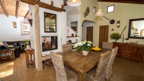 Een gezellig eetgedeelte met een houten tafel en rieten stoelen, omgeven door een rustiek interieur met zichtbare houten balken en een open haard.