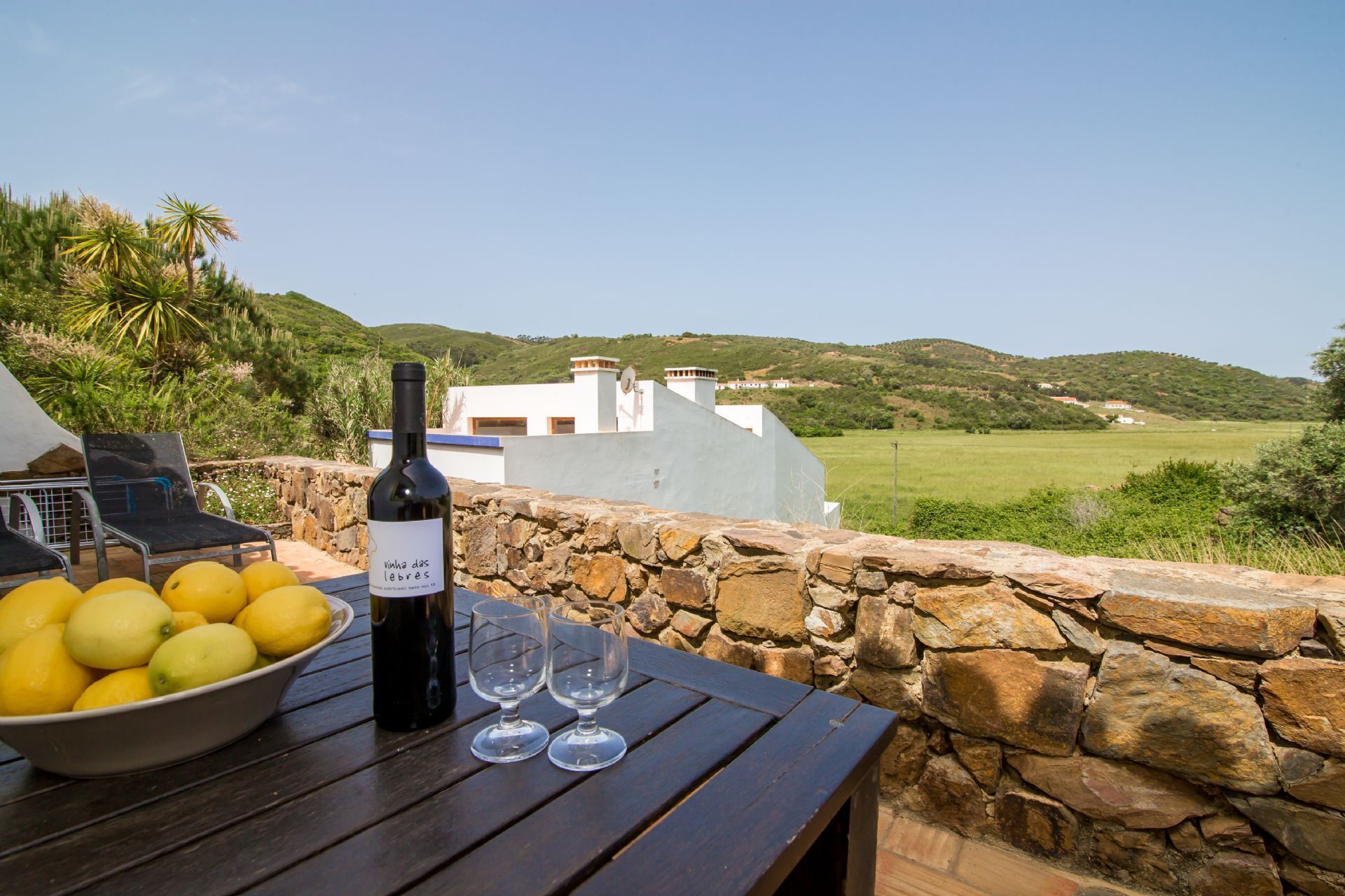 A table with a bottle of wine, glasses, and lemons on a wooden table with a stone wall backdrop.