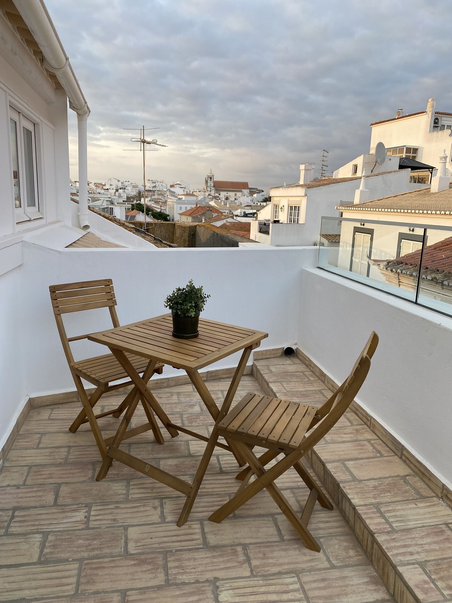 A balcony with a wooden table and chairs, a potted plant, and a view of a cityscape.