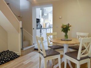 A dining area with a round table, four chairs, and a vase with flowers. A staircase is visible in the background leading to a kitchen area.