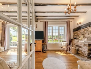 A room with a stone fireplace, wooden flooring, a large window with curtains, and a chandelier.
