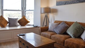 A living room with a brown leather sofa, a wooden coffee table, and a window with two cushions on the sill.