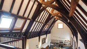 A high-ceilinged room with exposed wooden beams, a staircase with a wooden handrail, and a dining area with a blue sofa and a table set for four.