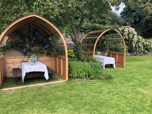Twee houten bogen met tafels en stoelen, omgeven door groen en bomen.