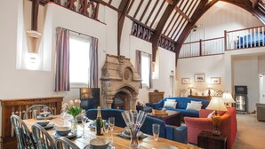 A dining area with a wooden table set for a meal, a stone fireplace, and a blue sofa in a spacious room with high ceilings and exposed beams.