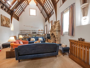 Un loft spacieux avec de hauts plafonds, des poutres apparentes, une cheminée en pierre et un coin salon confortable avec un canapé bleu et des fauteuils.