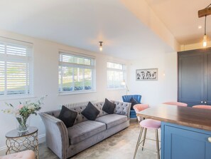 Una sala de estar moderna con un sofá gris, una silla azul, una mesa de comedor de madera y una zona de cocina con un armario azul.