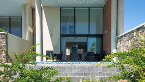 Une entrée d’hôtel moderne avec une façade en verre, un espace extérieur aménagé pour s’asseoir et un mur en pierre.