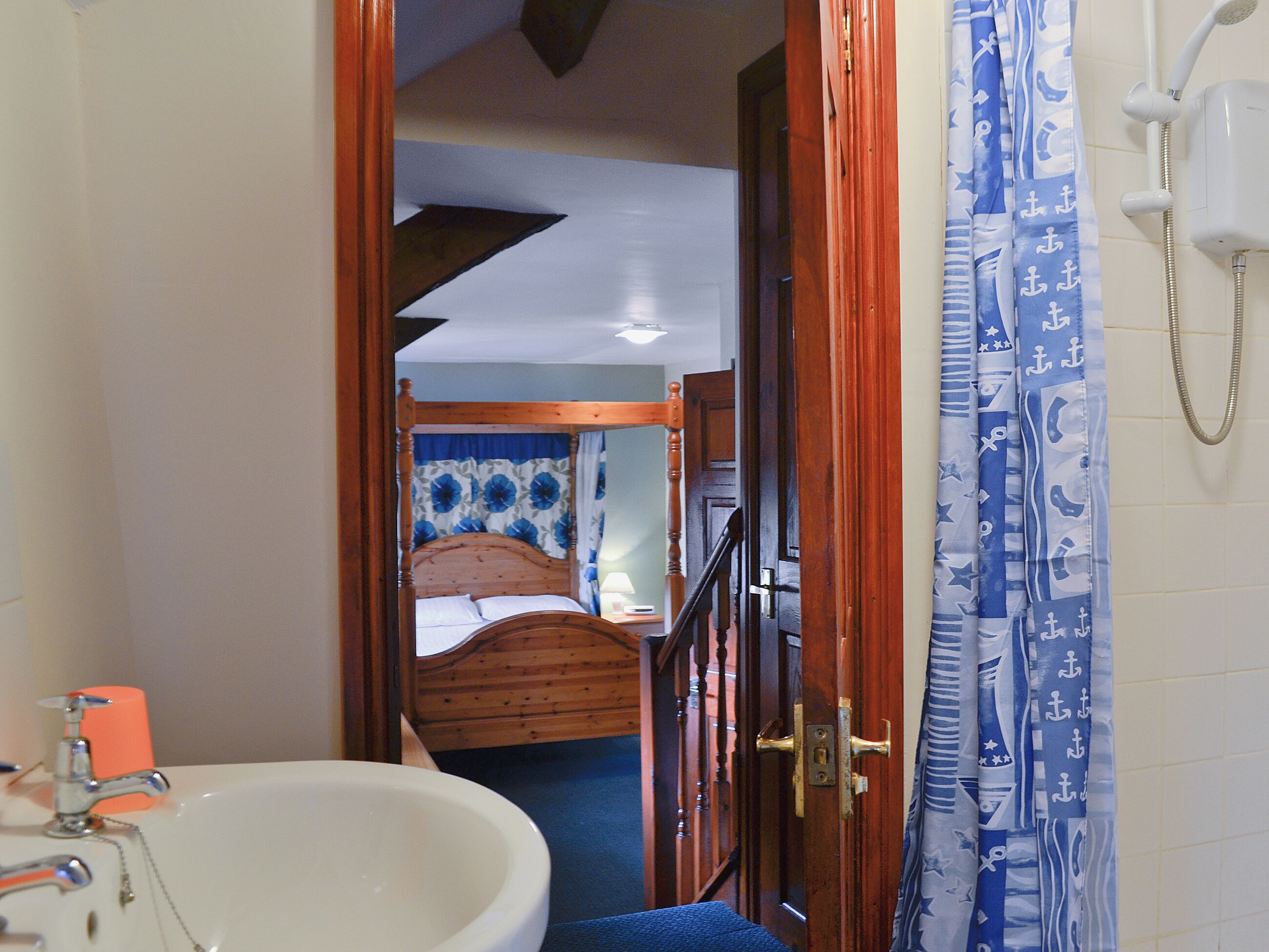 A bathroom with a sink, shower, and wooden door leading to a bedroom with a bed and curtains.