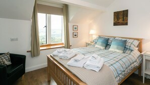 A bedroom with a large bed, a nightstand, a lamp, a window with curtains, a painting on the wall, and a wooden floor.
